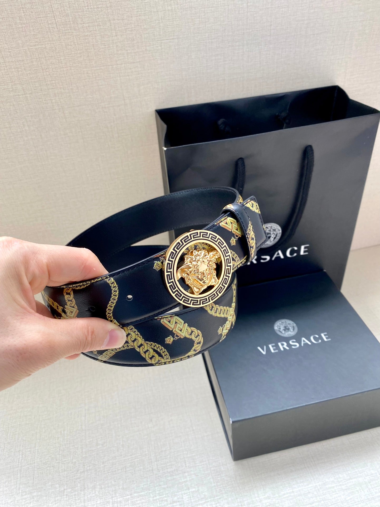 Hand holding navy Versace belt with gold pattern and round medallion buckle