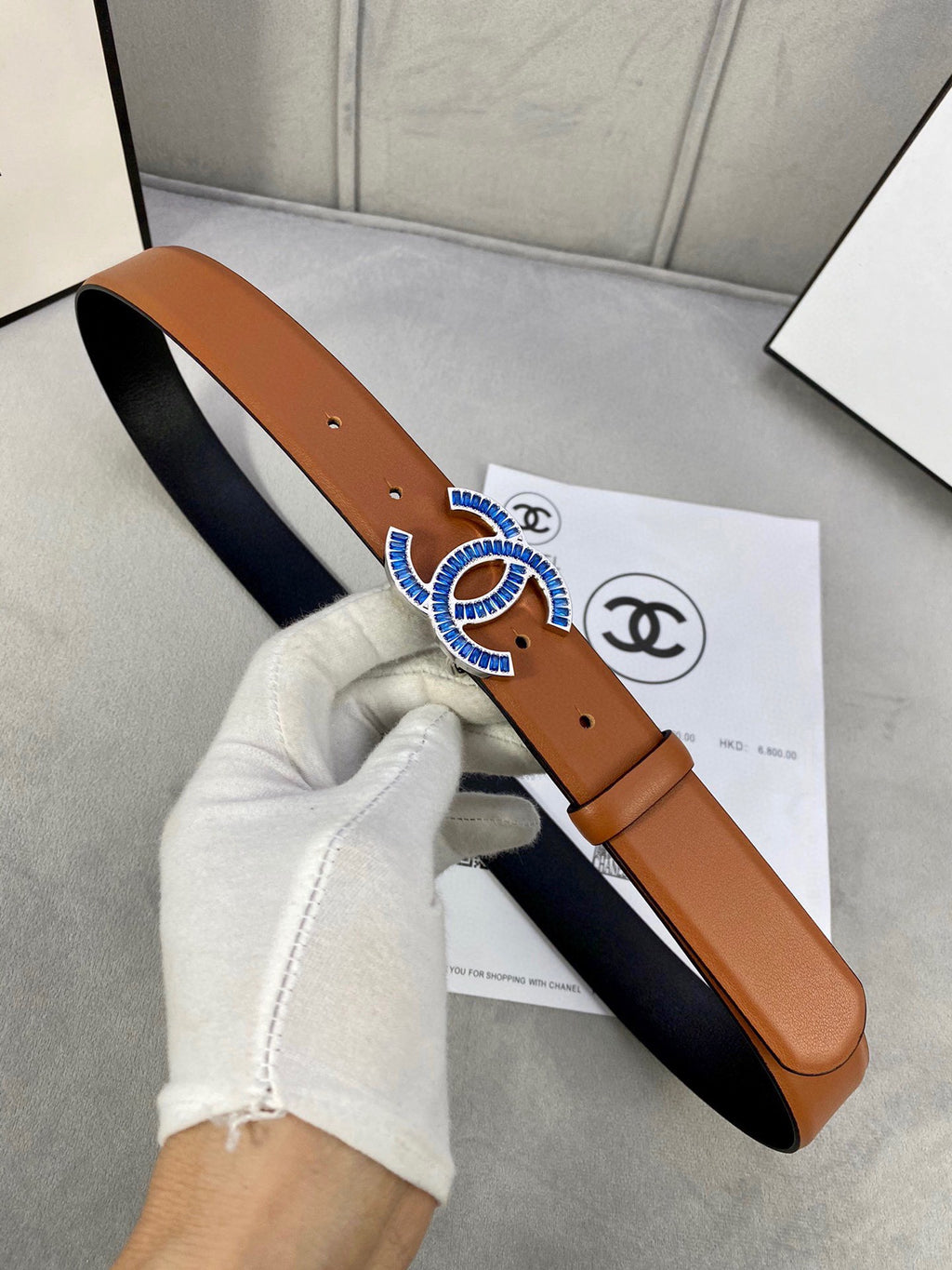 Tan leather belt held up, multicolor jeweled logo buckle centered