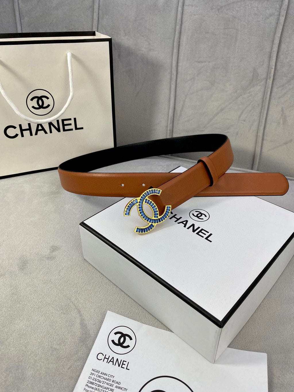 Tan leather belt on box and papers, featuring multicolor jeweled logo buckle