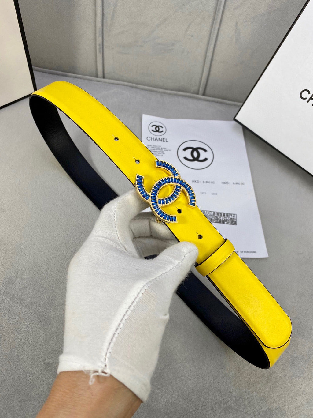 Chanel belt held by white glove, yellow strap with blue CC buckle and black underside
