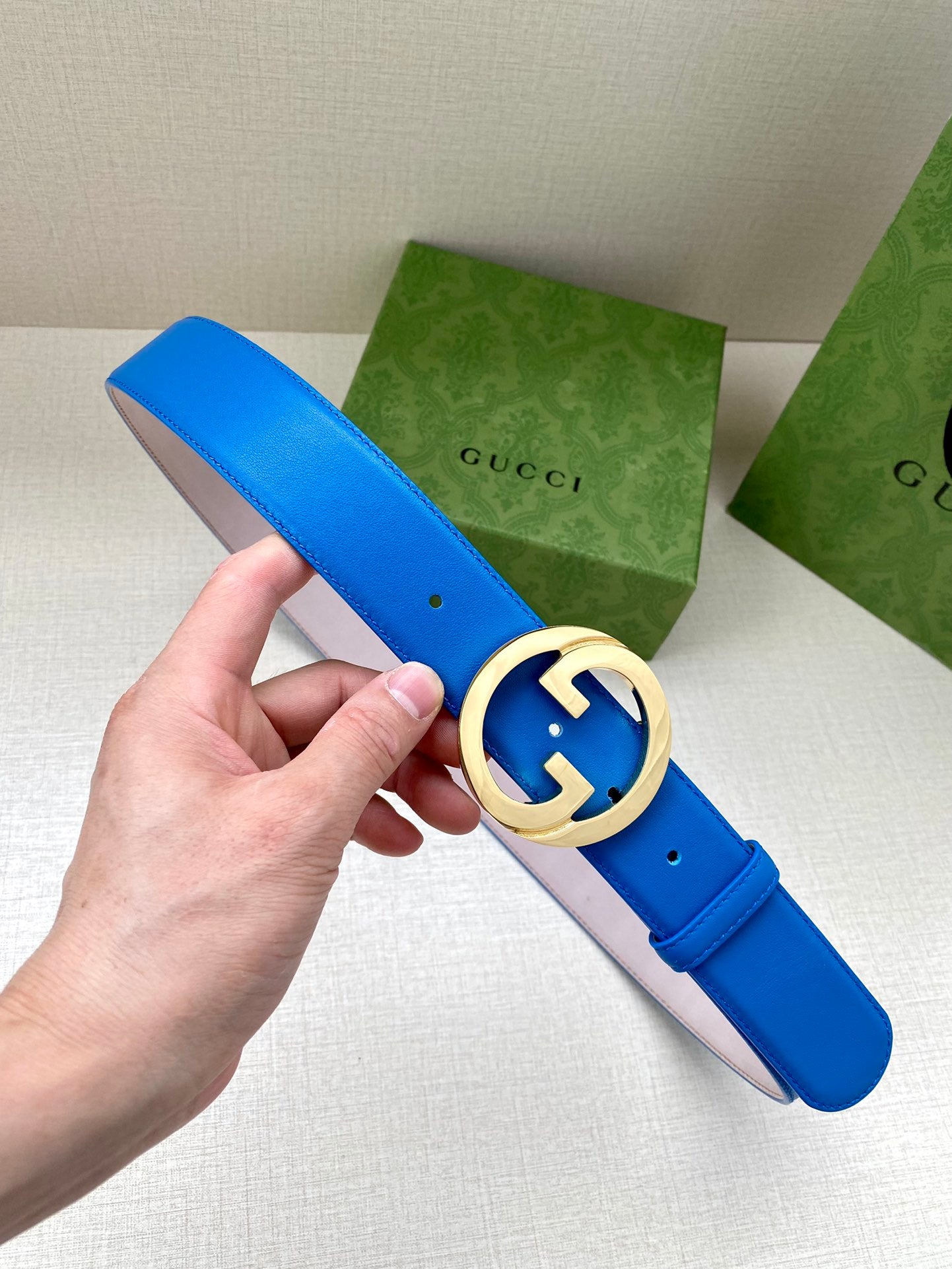 Blue Gucci belt with gold-tone GG buckle in round frame, pin holes visible.