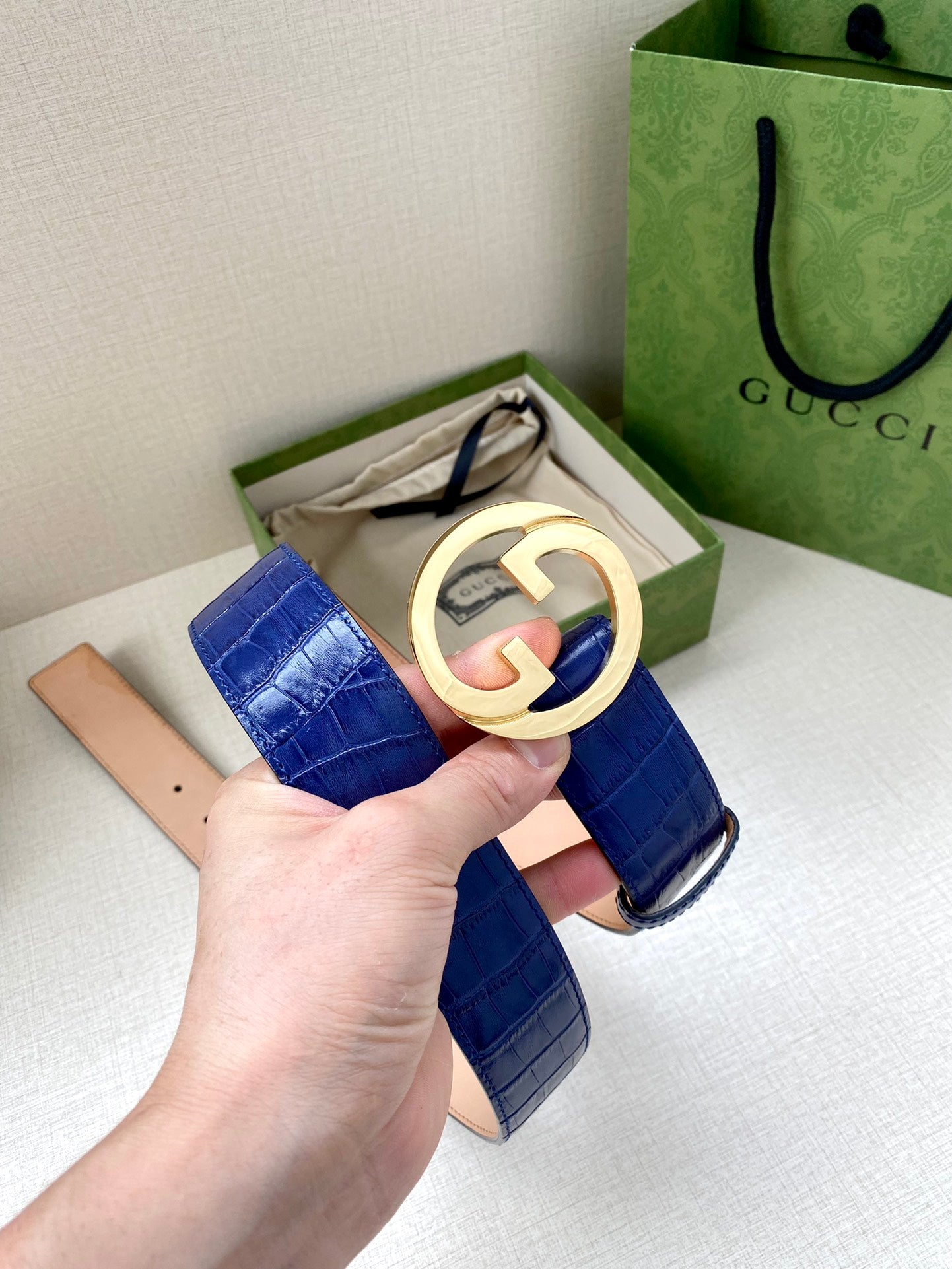 Blue croc-embossed Gucci belt with gold-tone GG buckle held in hand
