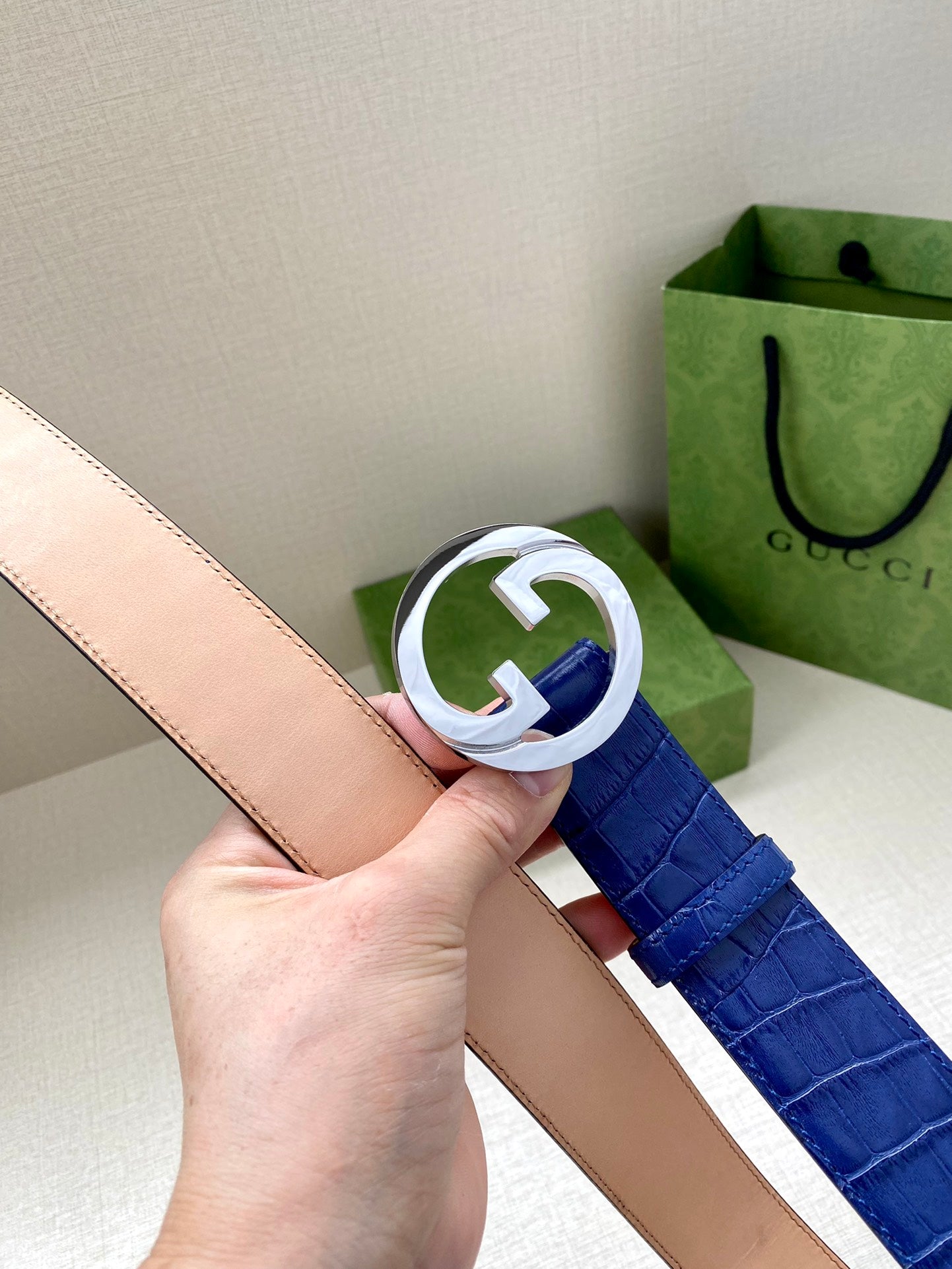 Blue croc-embossed Gucci belt with silver-tone GG buckle held over tan strap