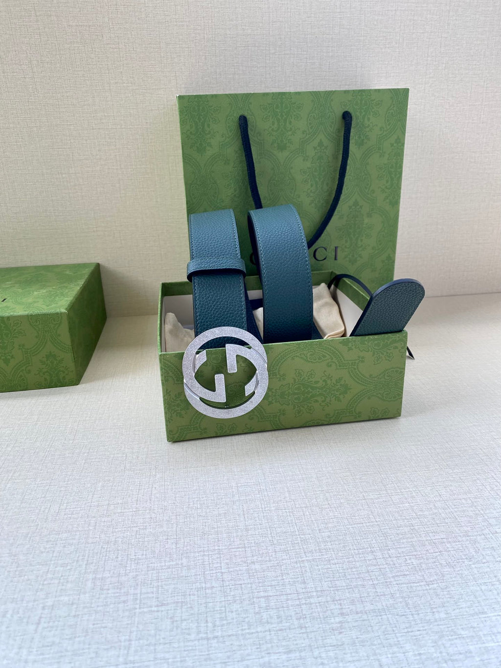 Teal leather belt with interlocking logo buckle, shown in green gift box