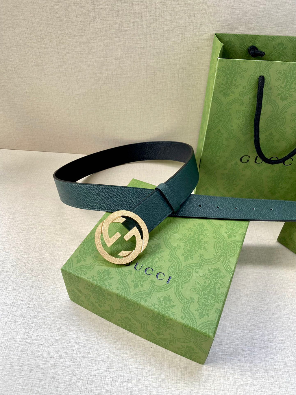 Teal leather belt with gold-tone interlocking logo buckle, resting on box