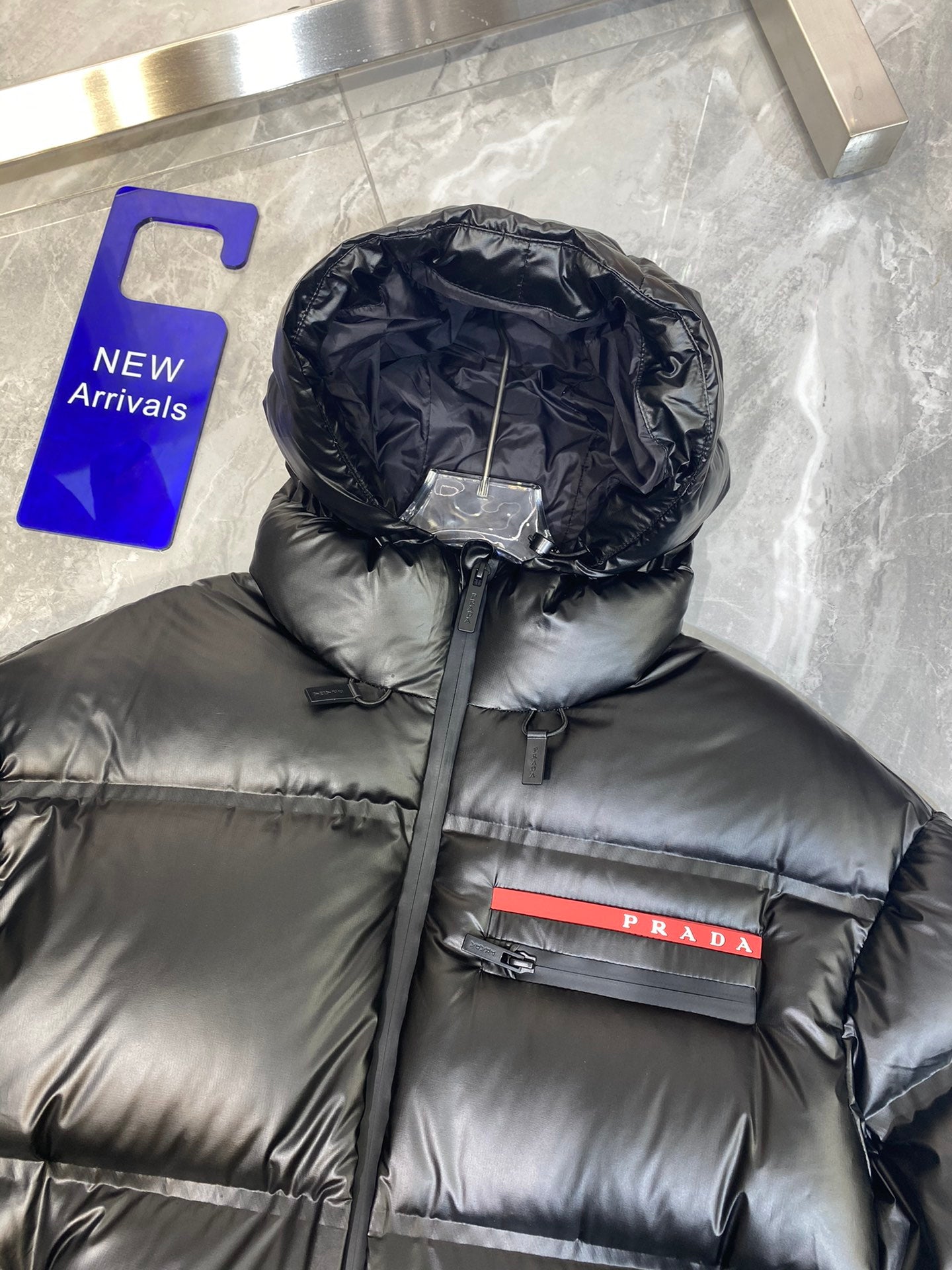 Black long puffer jacket with hood, zip front, and red Prada logo stripe detail