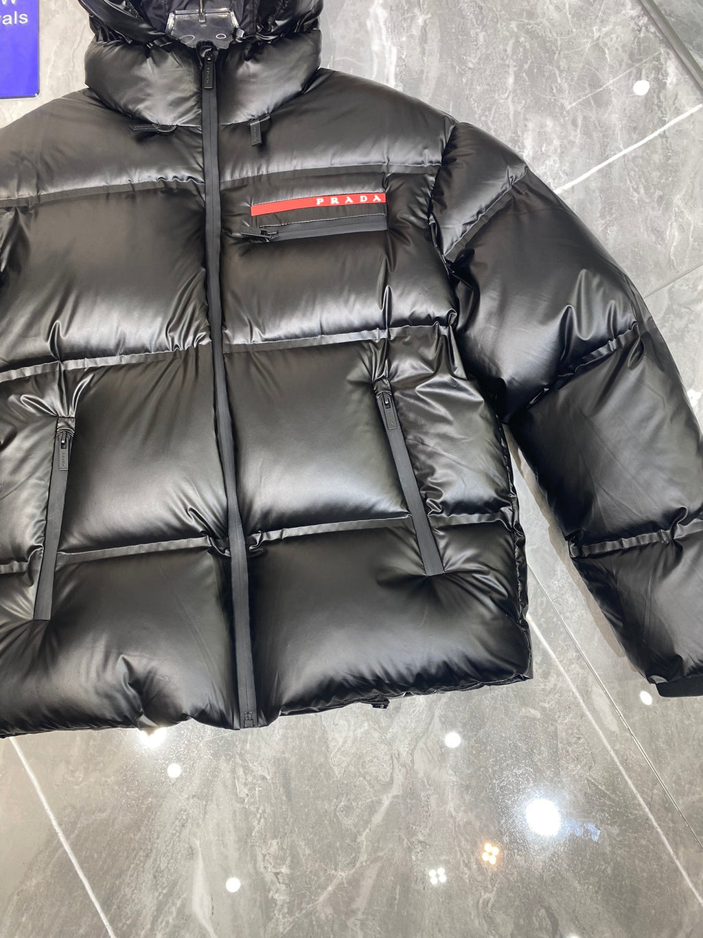 Black long puffer jacket with hood, front zip, quilted panels, and Prada logo stripe