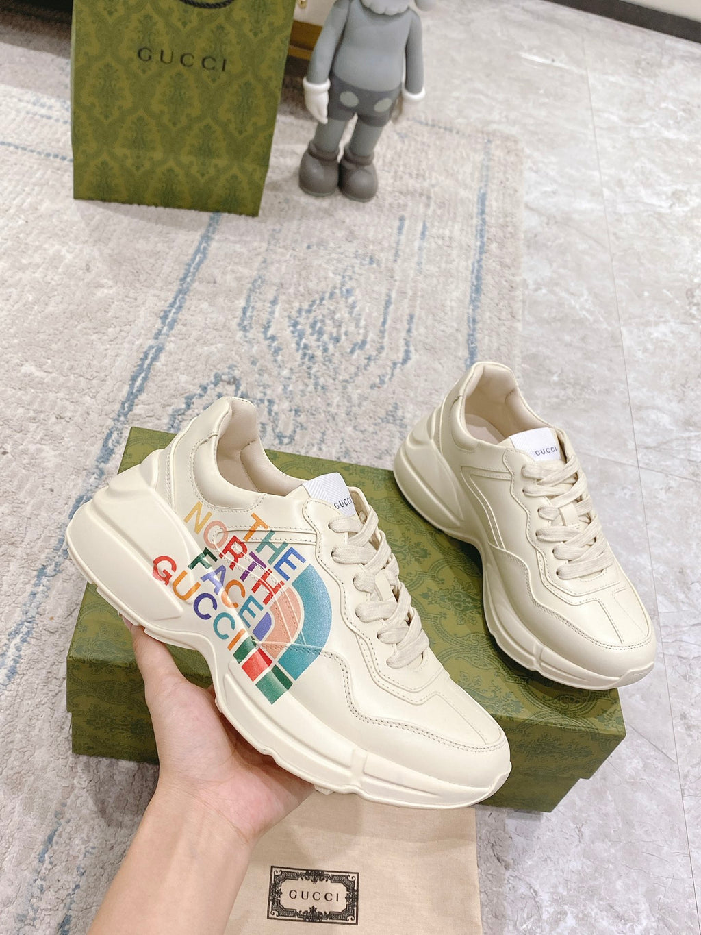 White lace-up sneakers held in hand, with The North Face Gucci multicolor logo