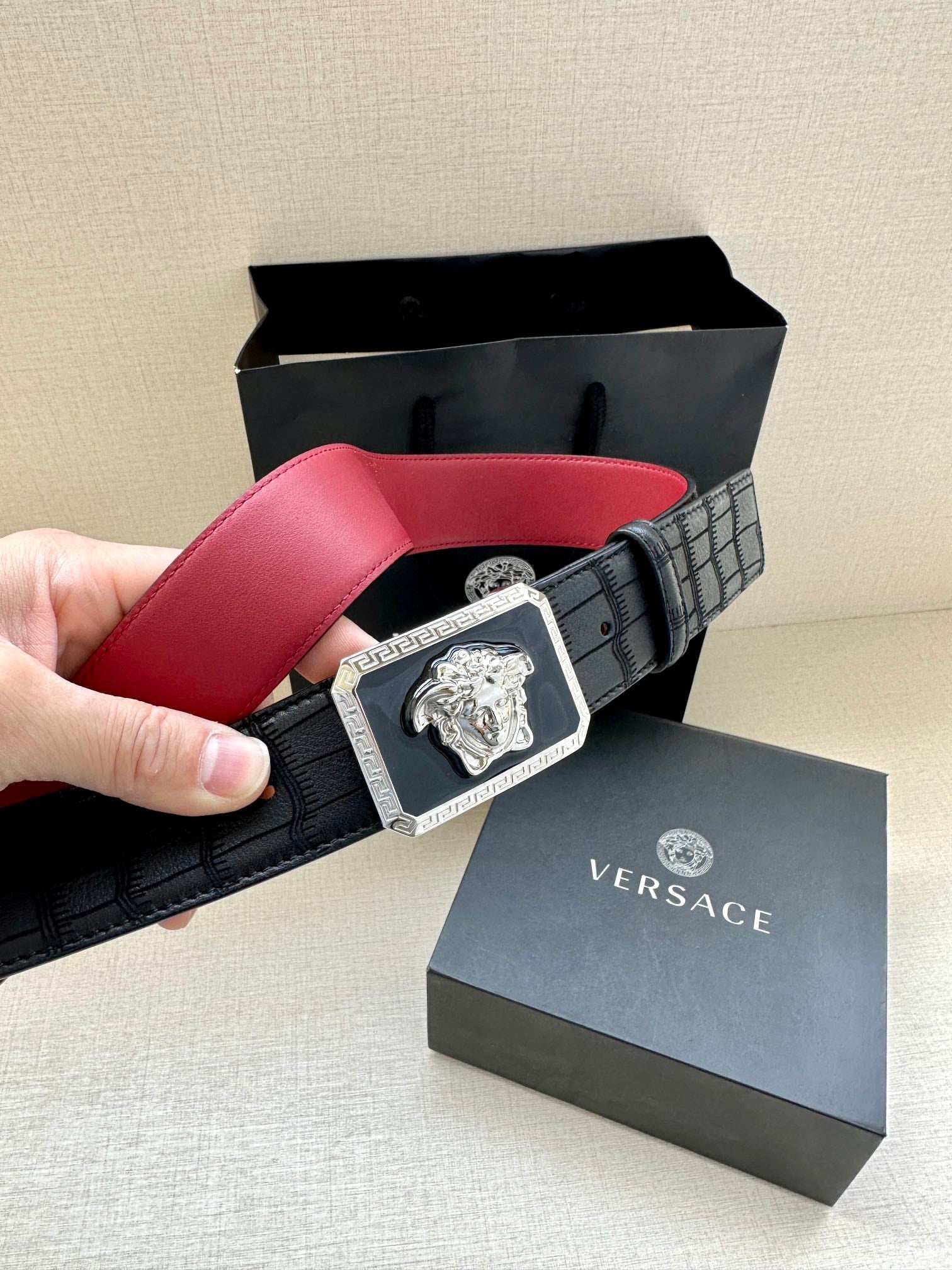Belt: black with red interior, square Medusa emblem buckle, held in hand.