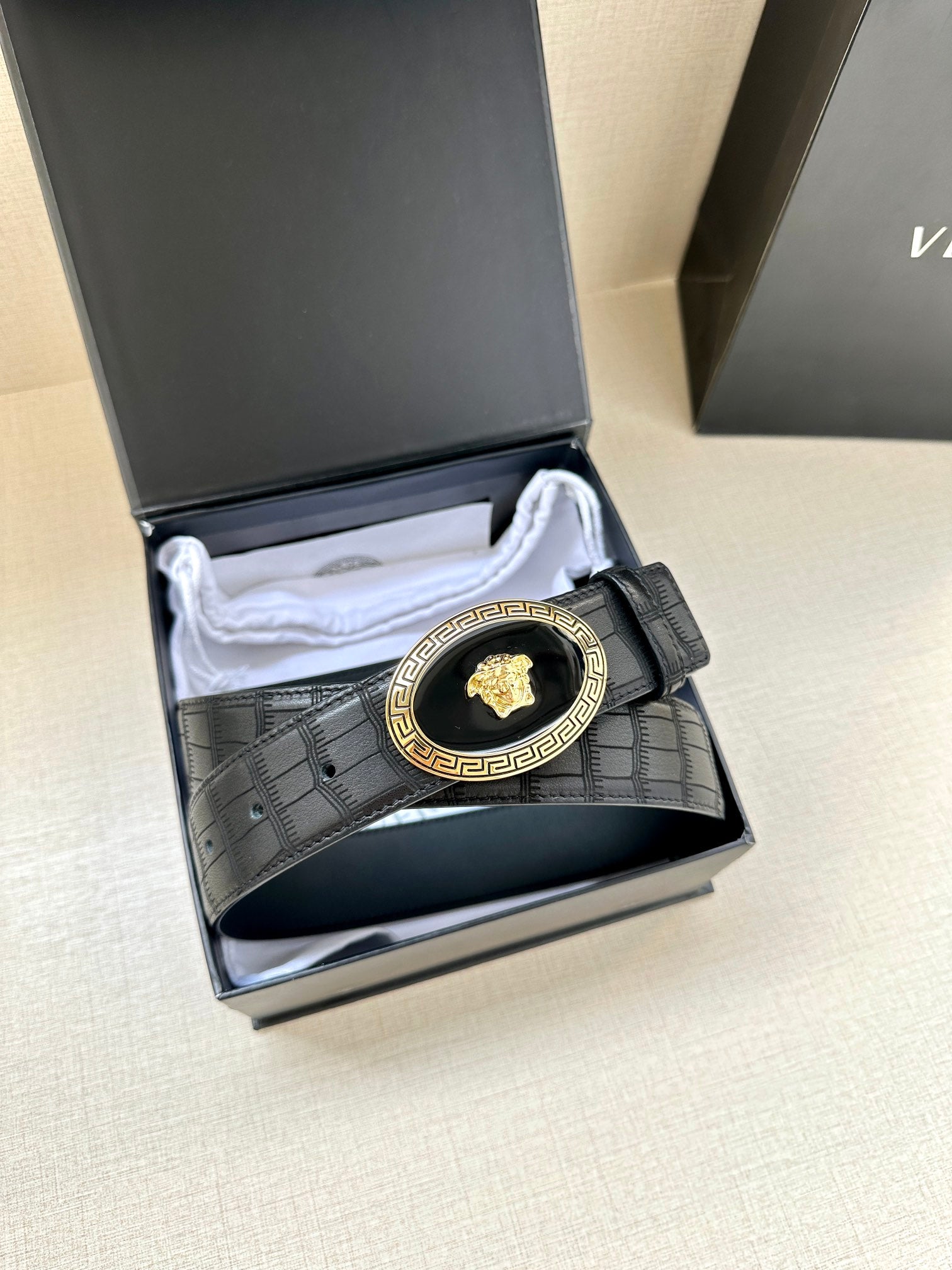 Black embossed leather belt with gold oval medallion buckle, laid inside box