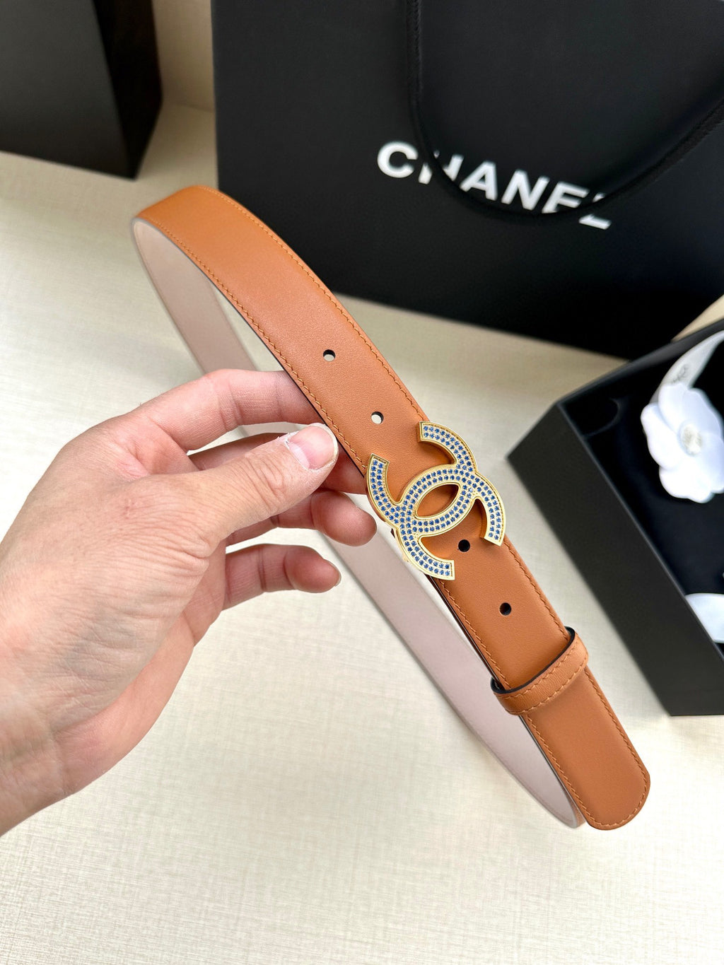 Tan leather belt with perforated holes and large silver-and-blue logo buckle