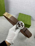 Brown leather belt with round silver G buckle, held in white-gloved hand
