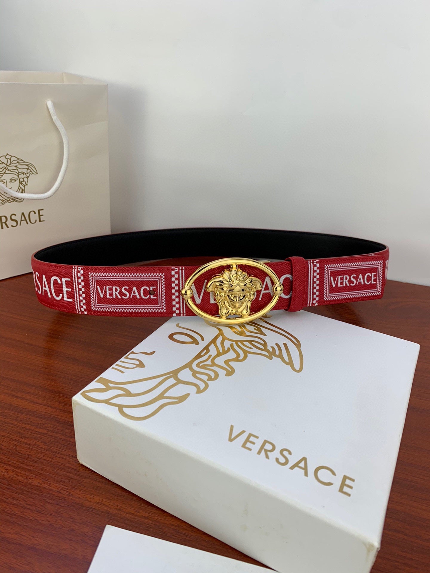 Versace belt on white box: red strap with repeating VERSACE text and gold Medusa buckle.