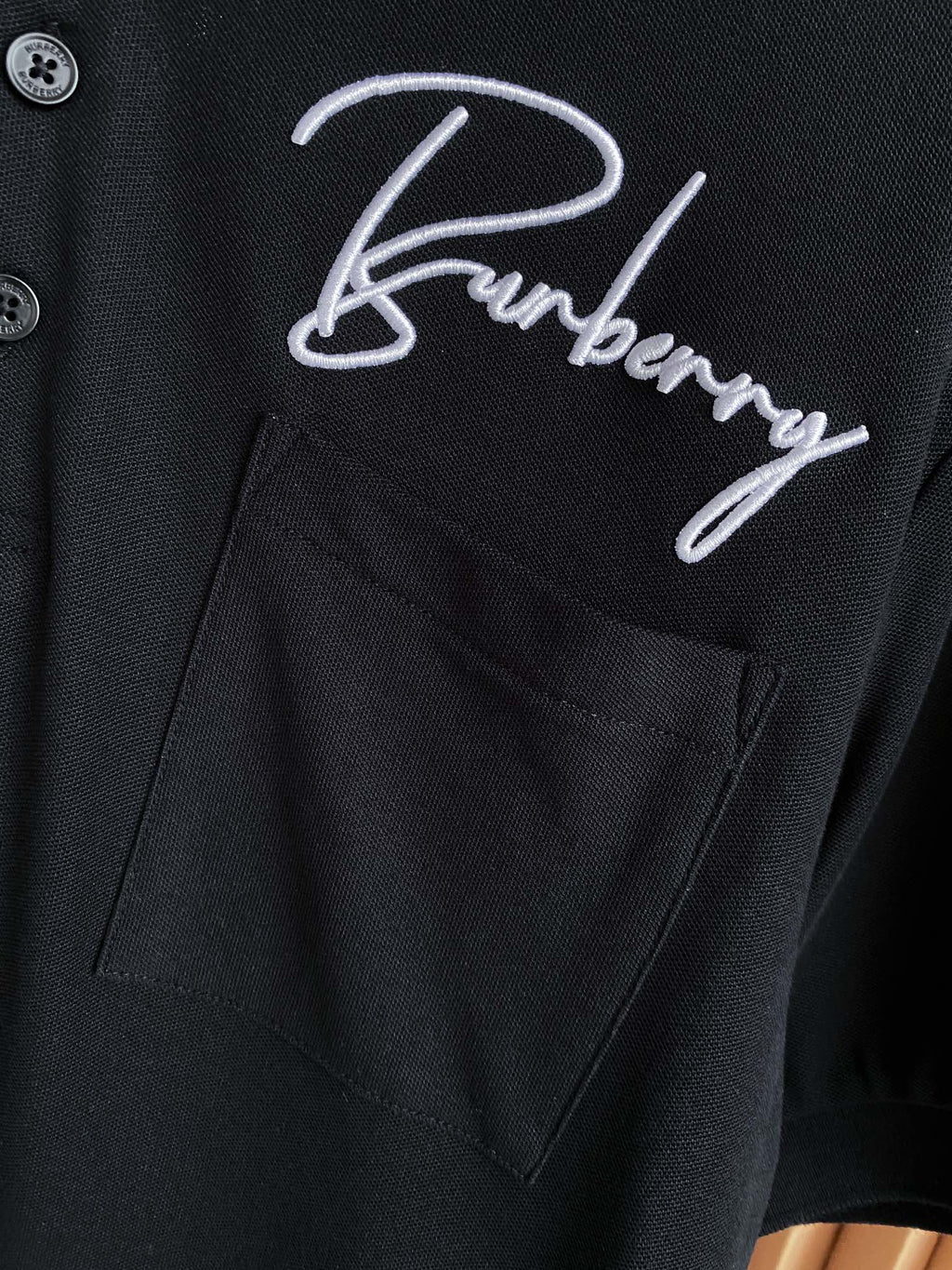 Black polo shirt pocket with white Burberry logo embroidery and button placket