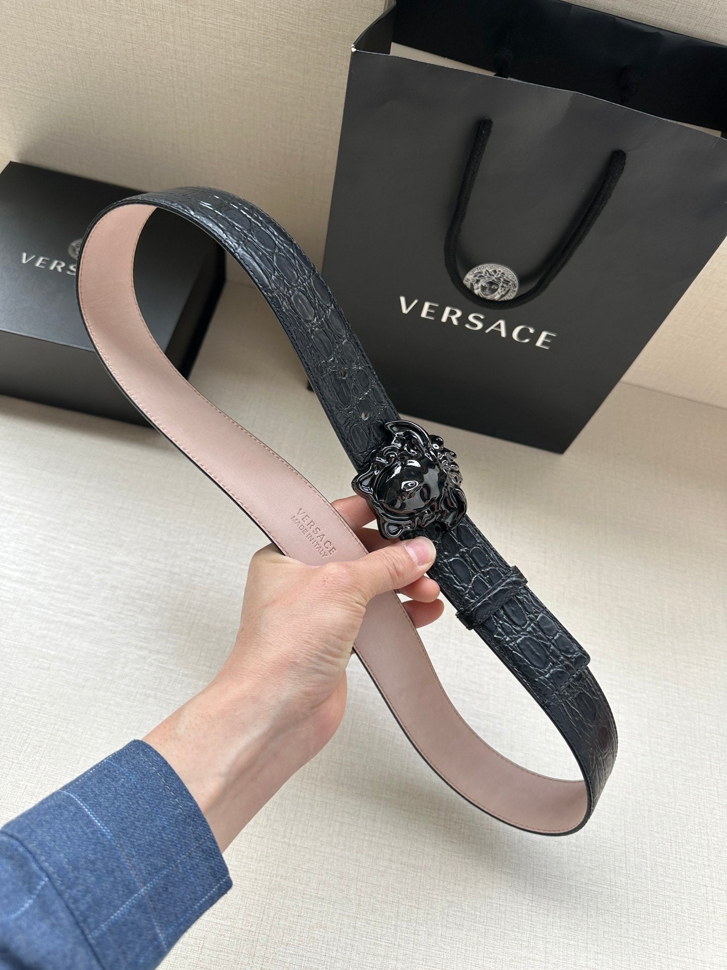 Black leather belt with croc texture and dark Medusa buckle detail, shown flat.