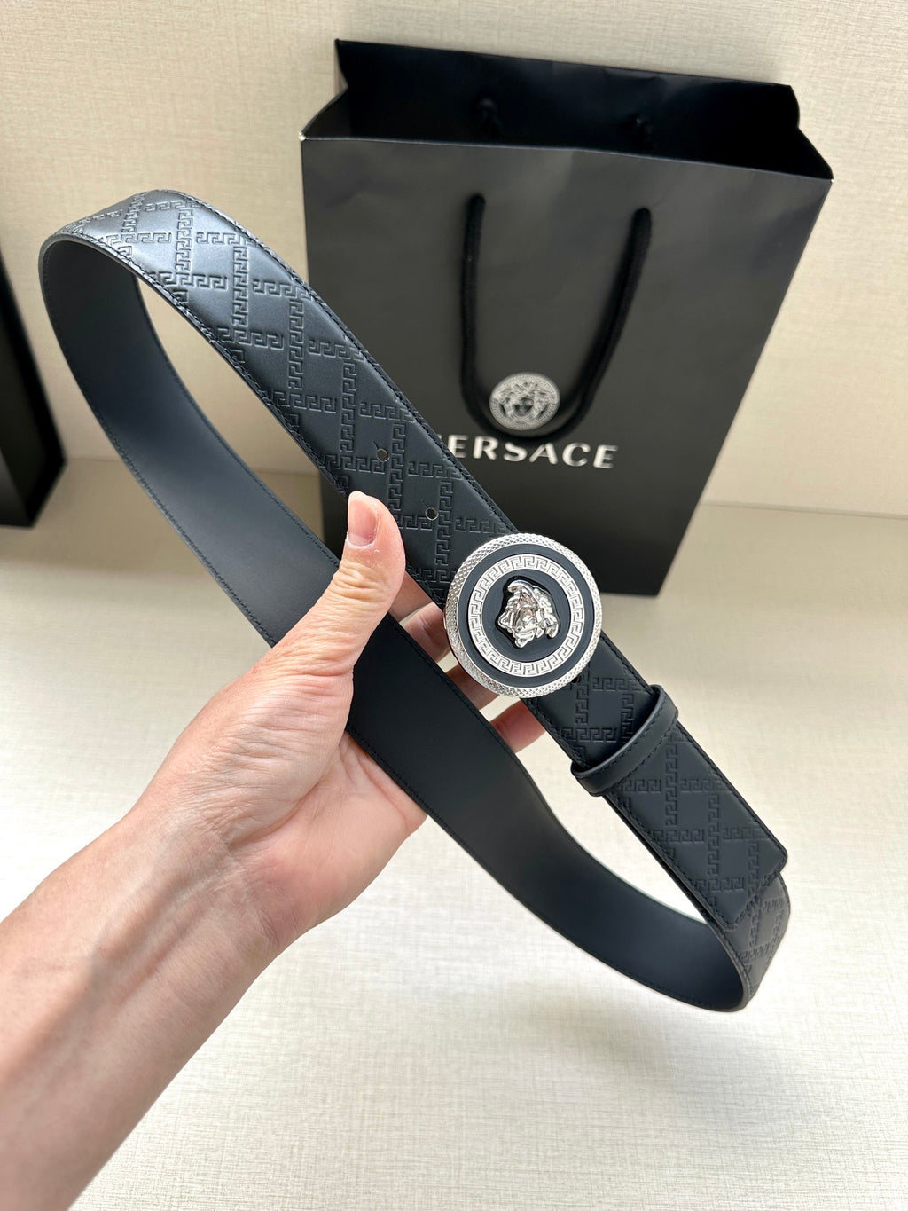 Versace belt held upright showing dark embossed strap and round medallion buckle
