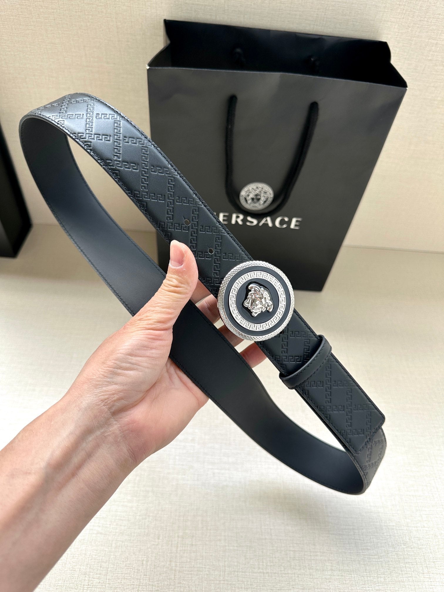 Versace belt held upright showing dark embossed strap and round medallion buckle