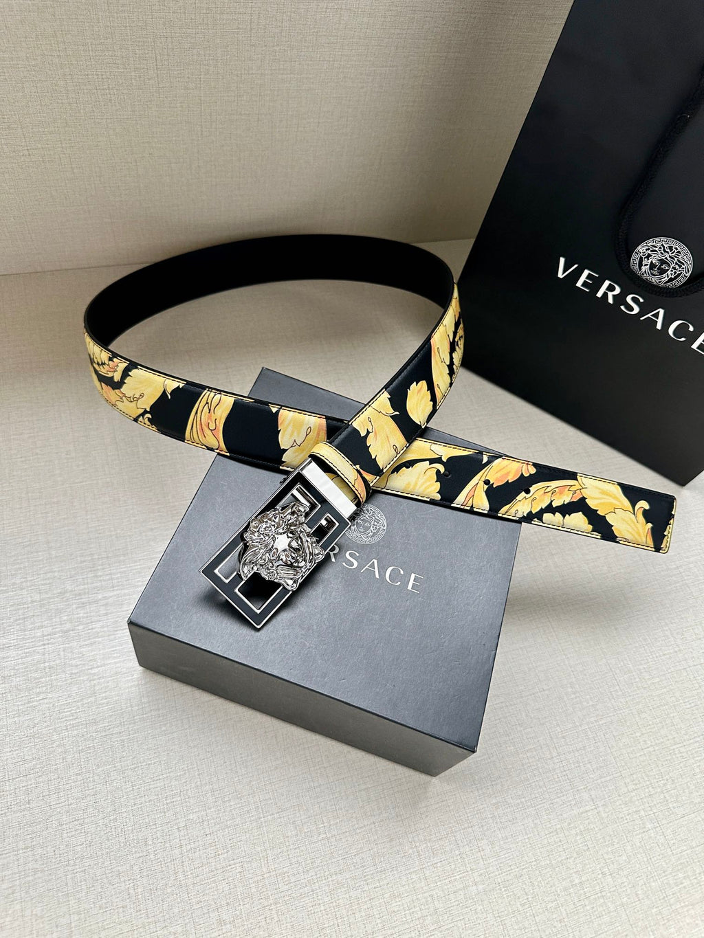 Versace belt laid flat with navy strap, gold floral print, and square buckle.