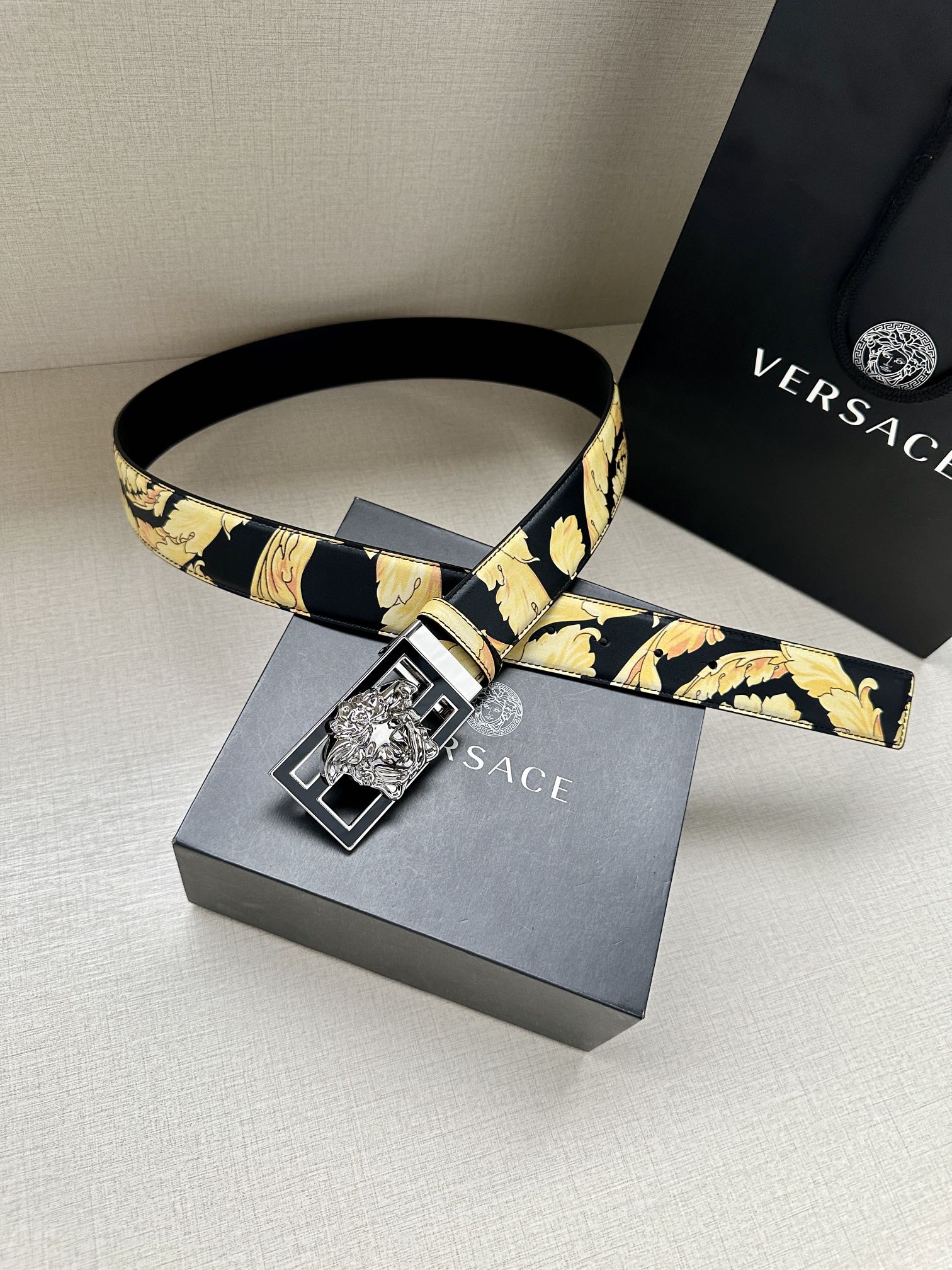 Versace belt laid flat with navy strap, gold floral print, and square buckle.