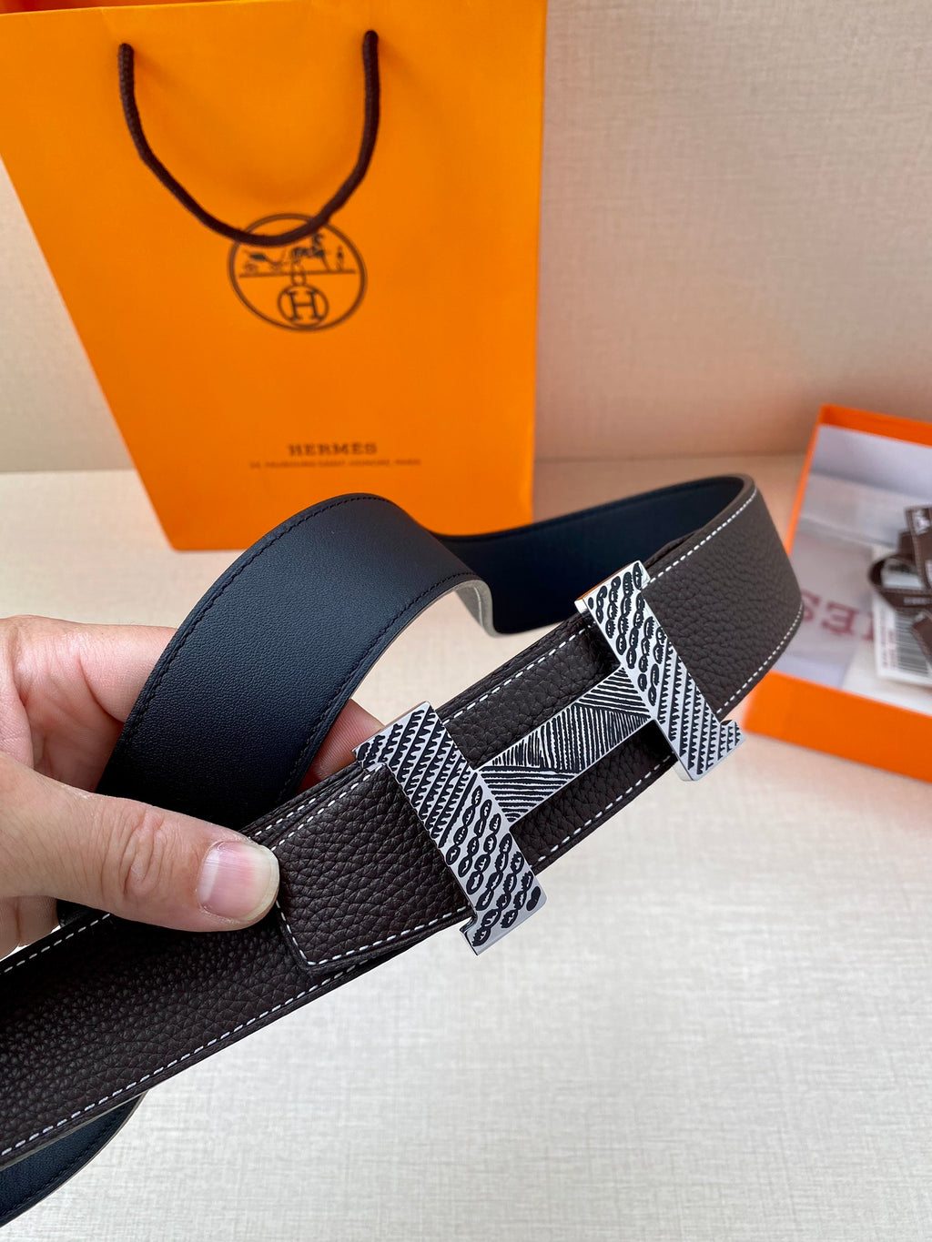 Hermes belt close-up on hand, dark leather with metal H buckle and patterned strap.
