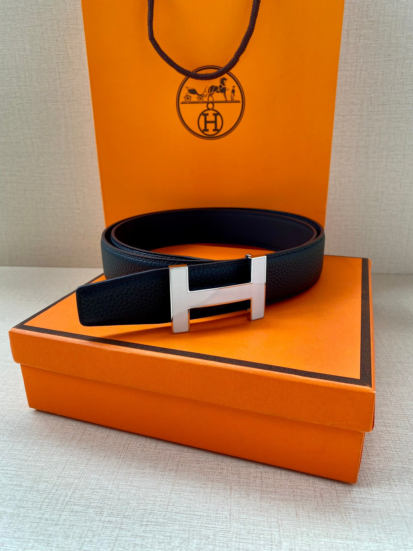 Dark blue leather belt with silver H buckle placed on orange box