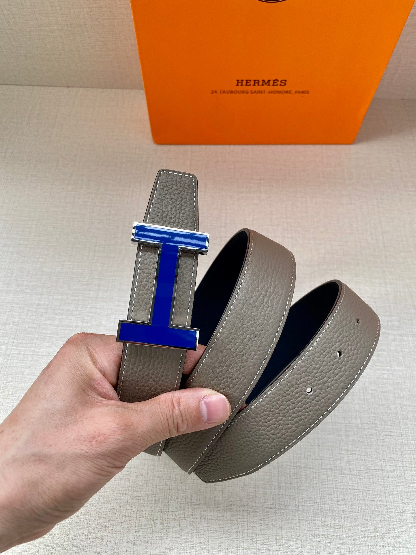 Two grey leather belts with blue enamel H buckles, one held in hand