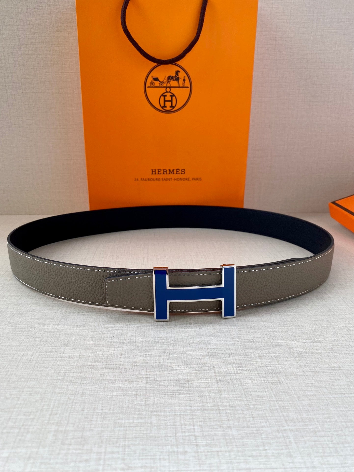 Grey leather belt with blue enamel H buckle, centered on the strap