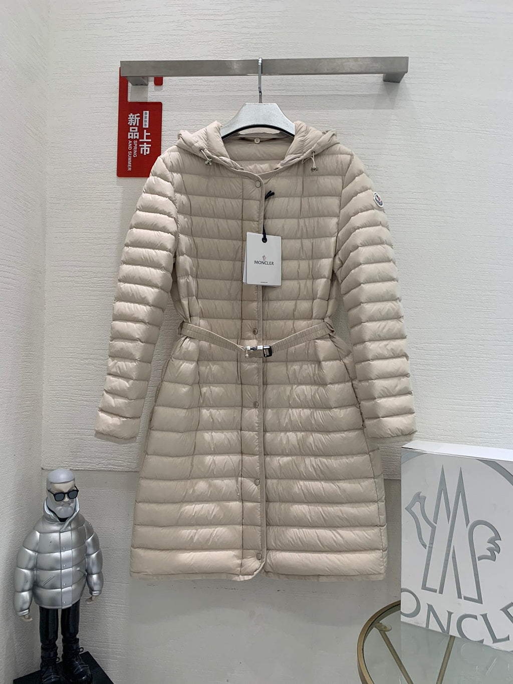 Long beige quilted puffer coat with hood, front zipper, and drawstring waist belt