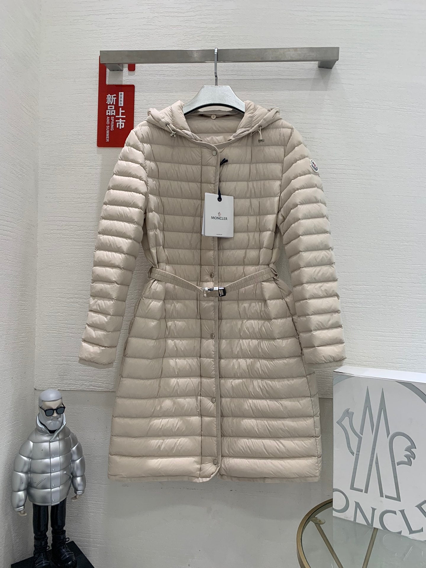 Long beige quilted puffer coat with hood, front zipper, and drawstring waist belt