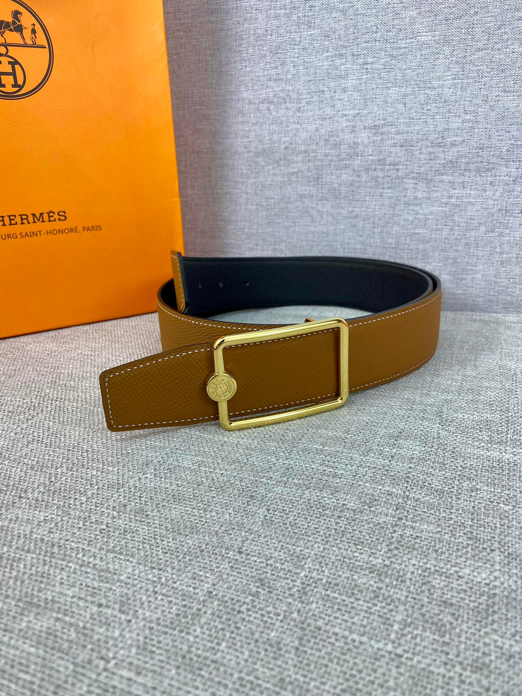 Tan leather belt with gold-tone square buckle and round logo charm on strap
