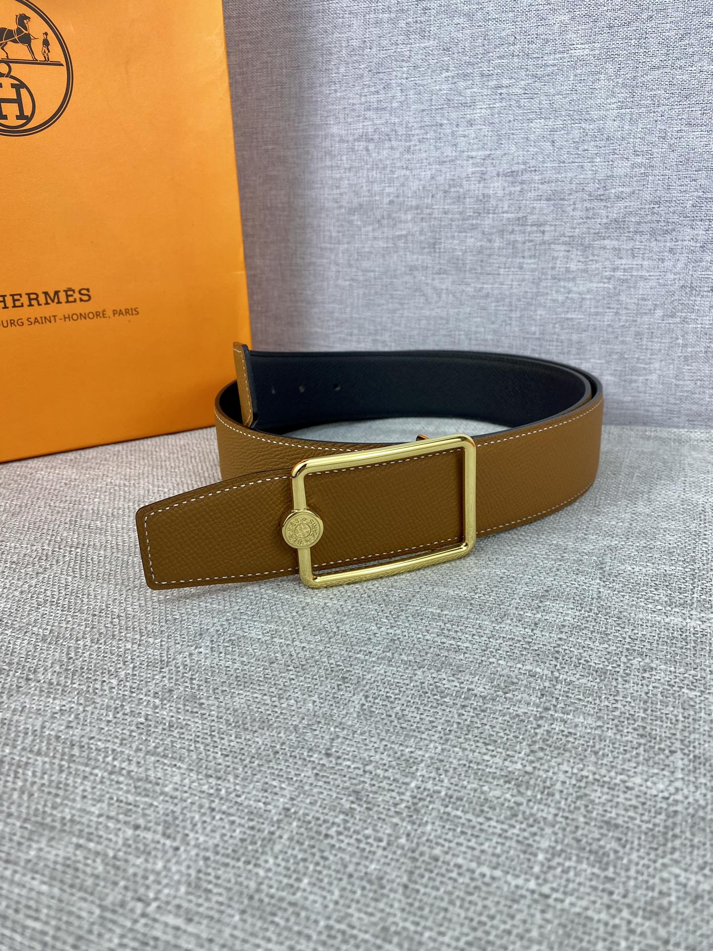 Tan leather belt with gold-tone square buckle and round logo charm on strap