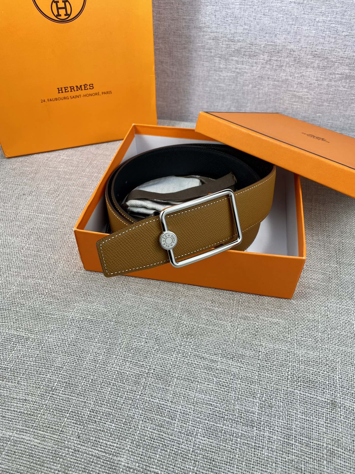Tan leather belt laid in box with gold-tone square buckle and logo button