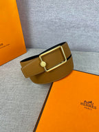 Tan leather belt with gold-tone square buckle and round logo plate, angled view