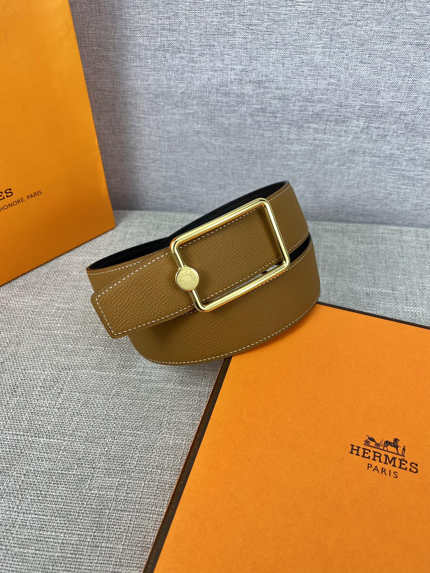 Tan leather belt with gold-tone square buckle and round logo plate, angled view