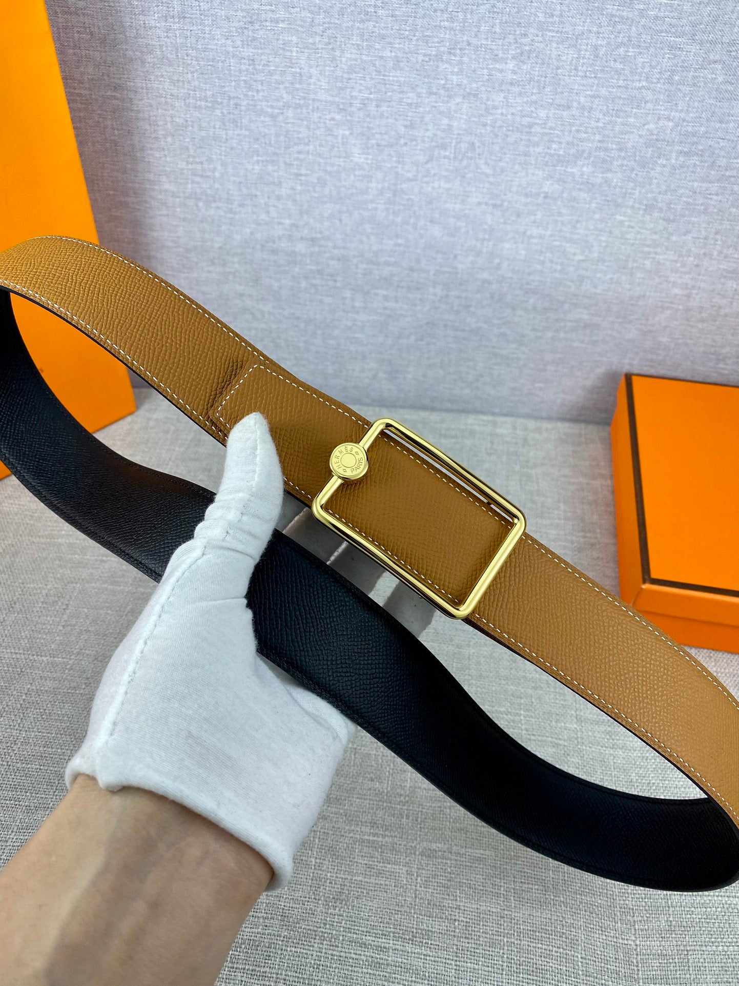 Tan leather belt with gold-tone square buckle over black backing, close-up