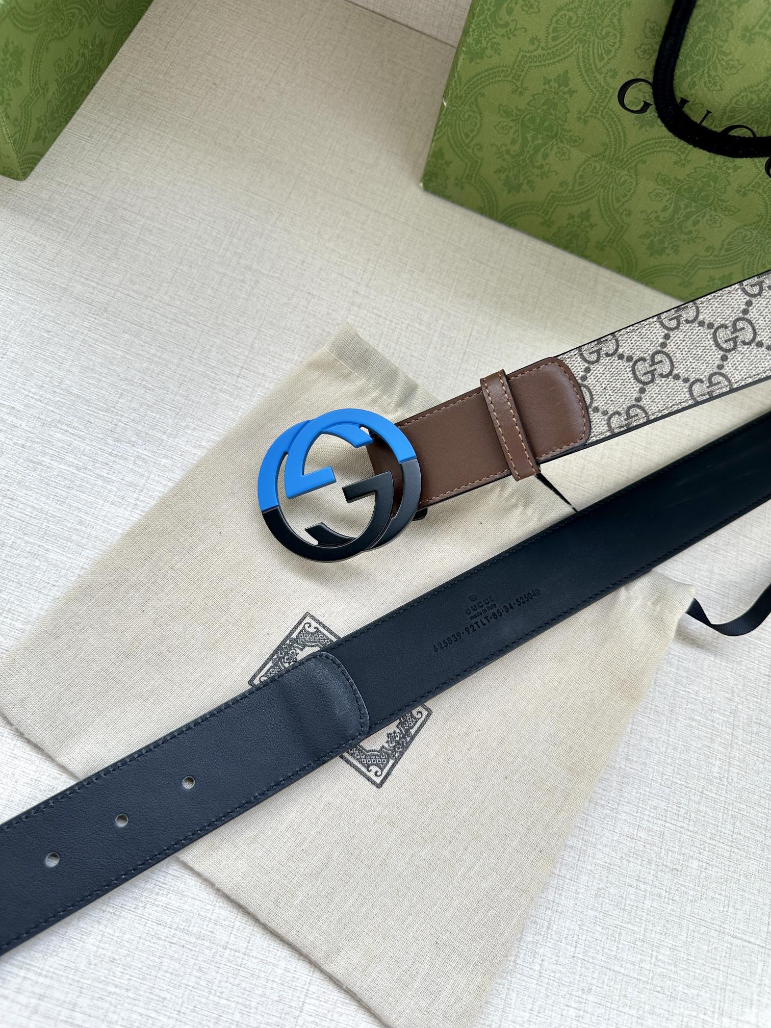 Brown leather belt with round black buckle beside dark navy strap on dust bag