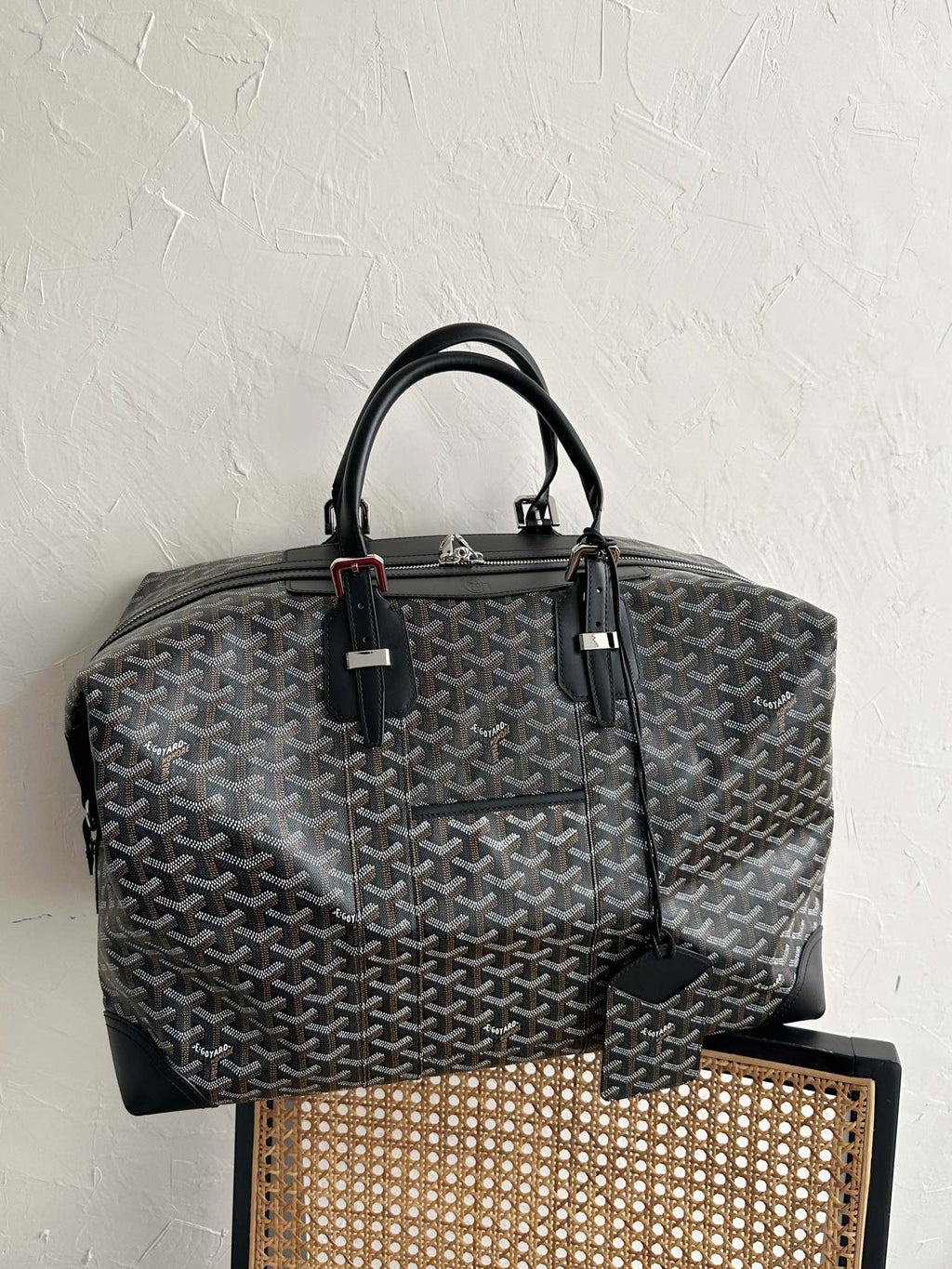 Tote bag with dark monogram pattern, black handles, and side leather strap detail.