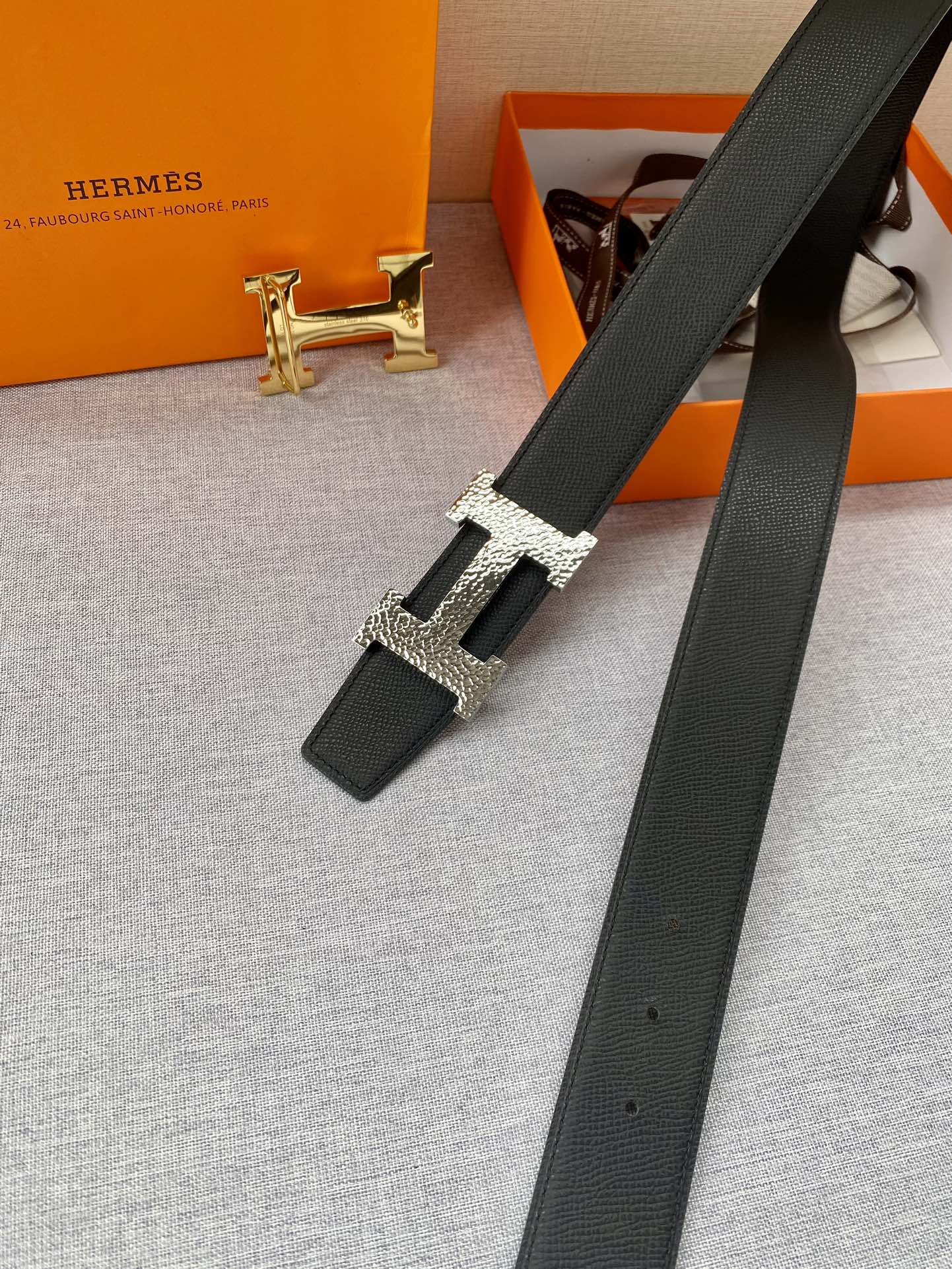 Black leather belt with gold H buckle and long strap, laid diagonally on fabric