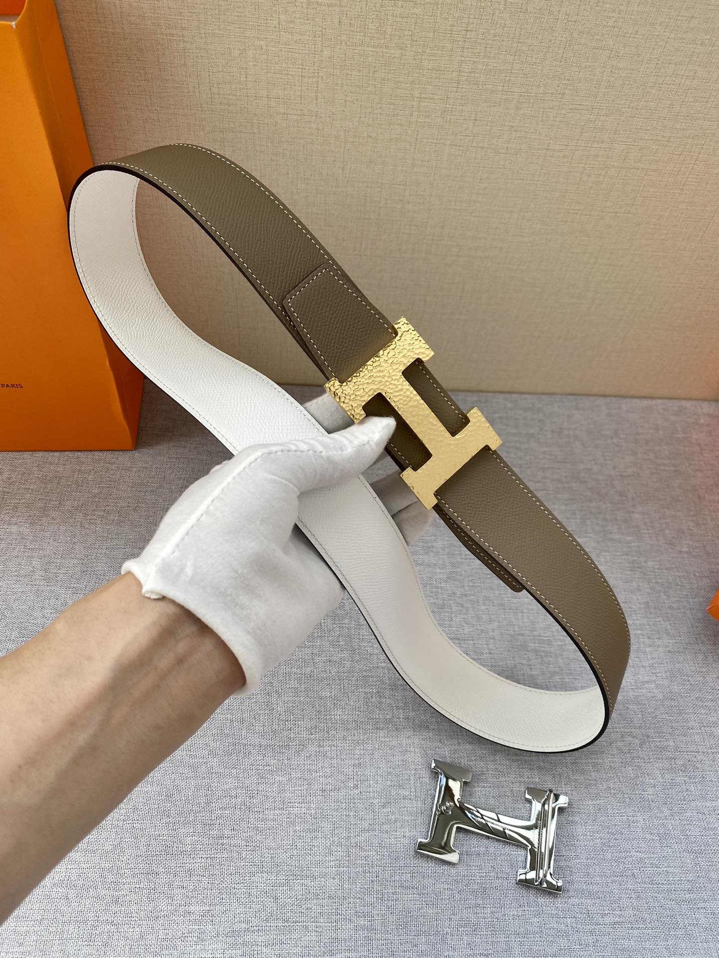 White-and-brown leather belt with gold-tone H buckle; outer strap shown curved.
