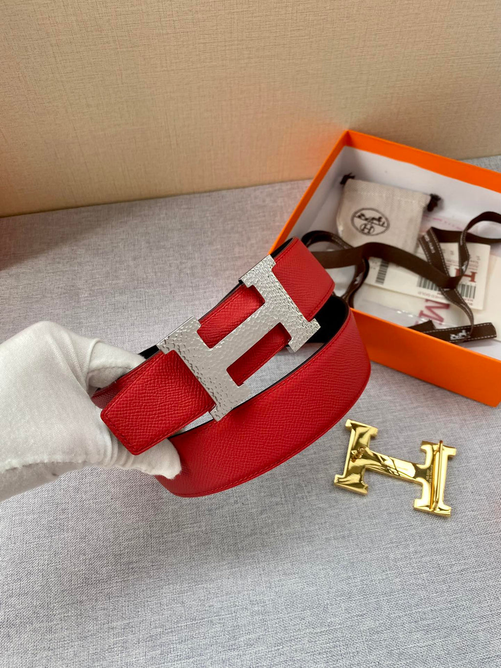 Red leather belt with silver-tone H buckle, resting on gray fabric with box
