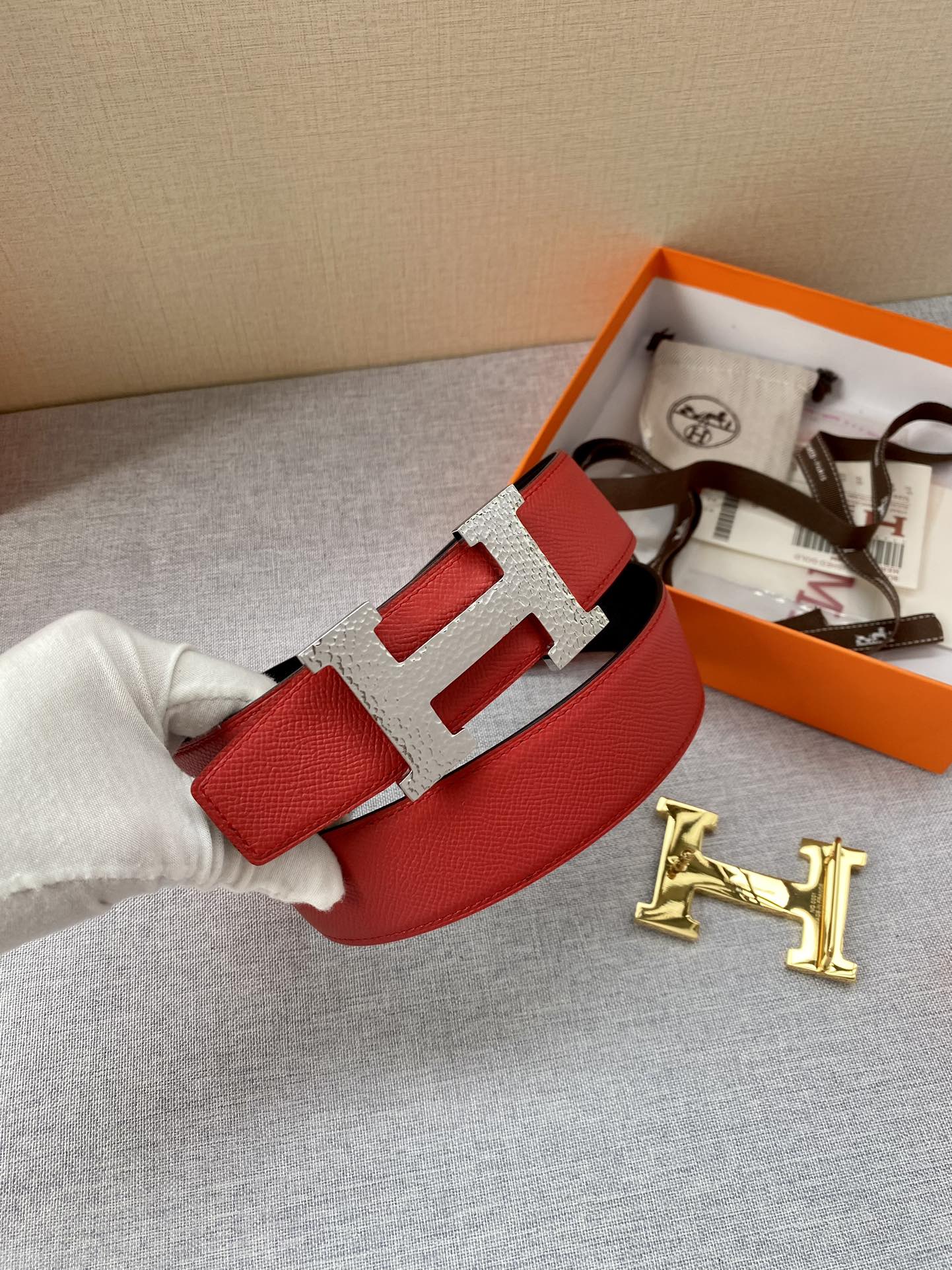 Red leather belt with silver-tone H buckle, laid on gray fabric with box contents