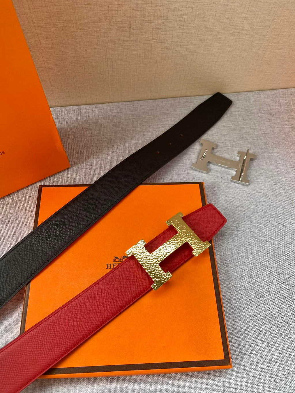 Red and black leather belt with gold-tone H buckle, laid on orange packaging