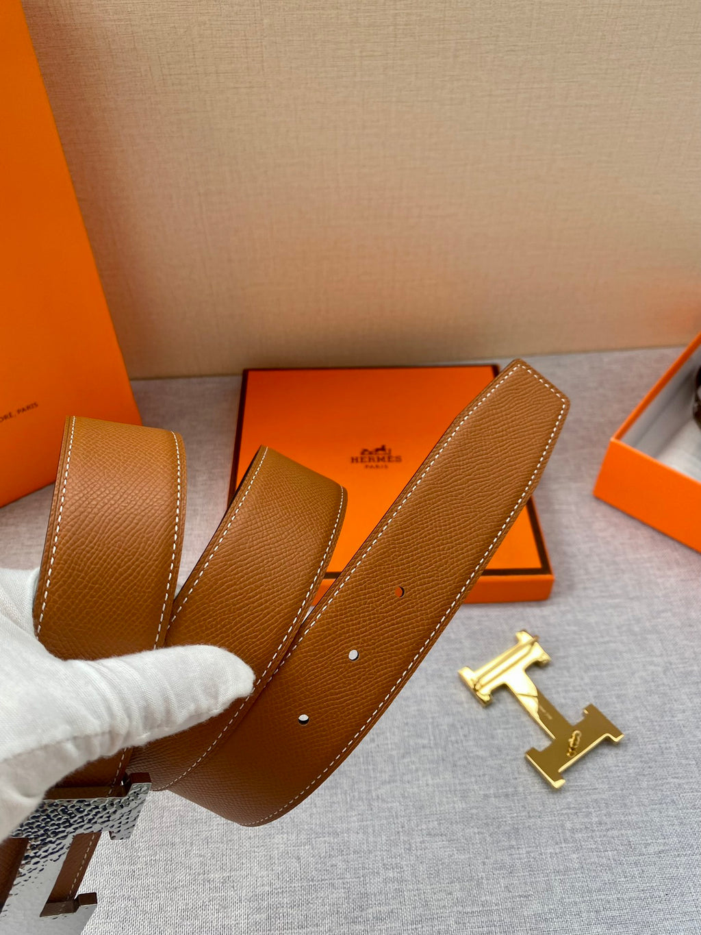 Tan leather belt strap with multiple holes and gold-tone hardware; packaging nearby.