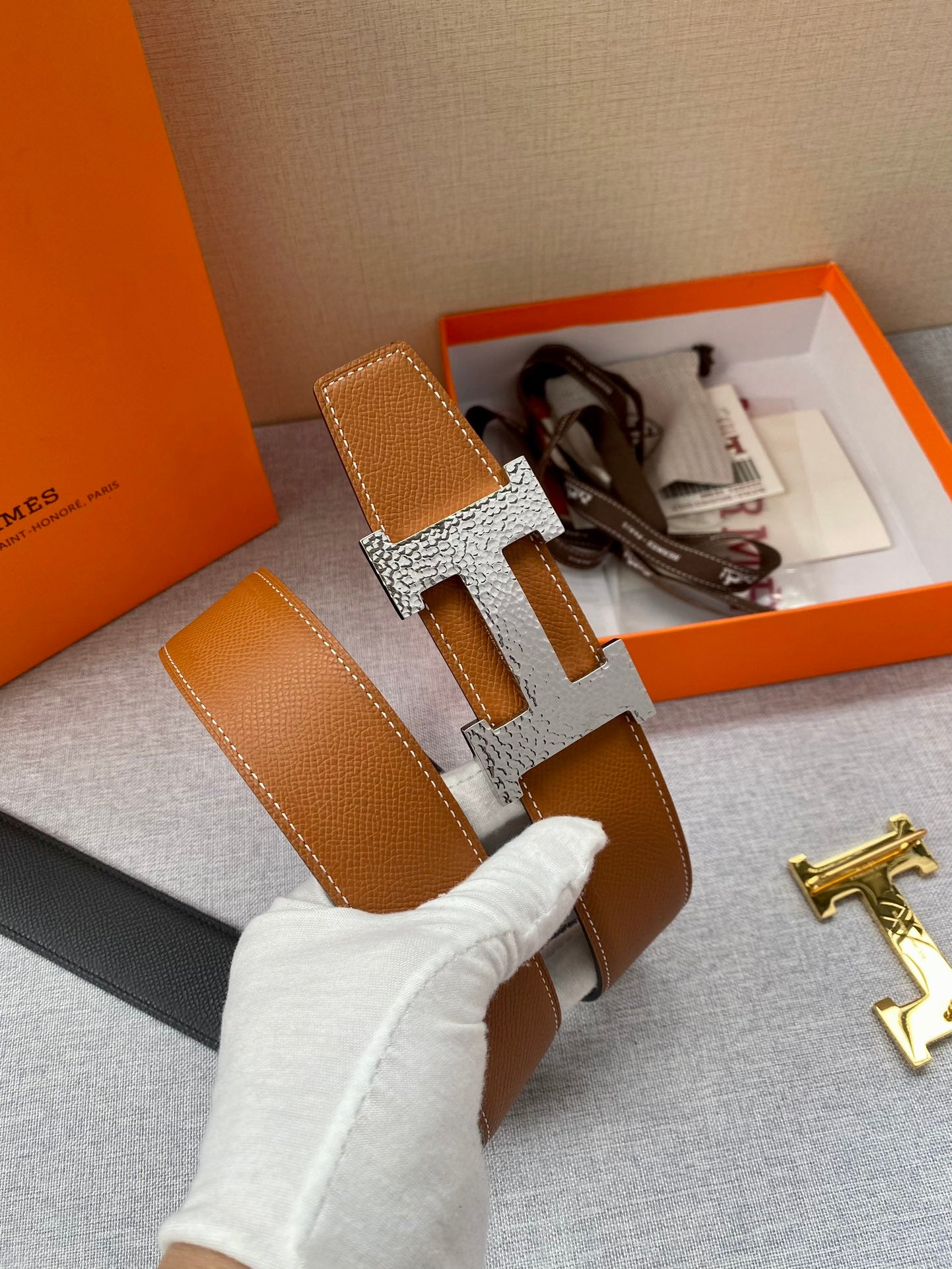 Tan leather belt with gold-tone H buckle and keeper; partial view of box insert.
