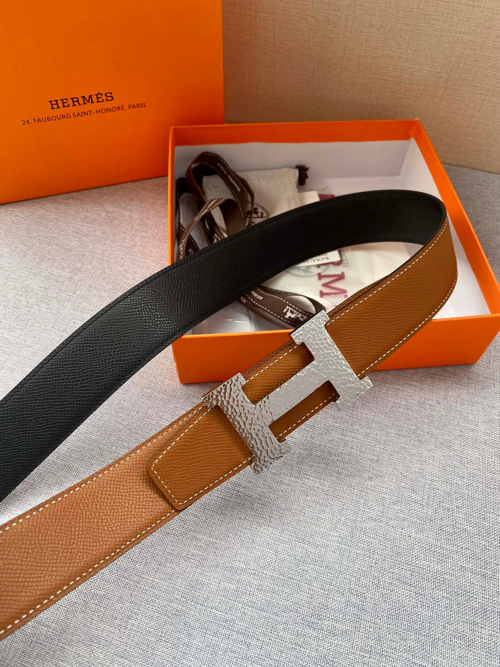 Tan leather belt with black strap back and gold-tone H buckle; side angle view.