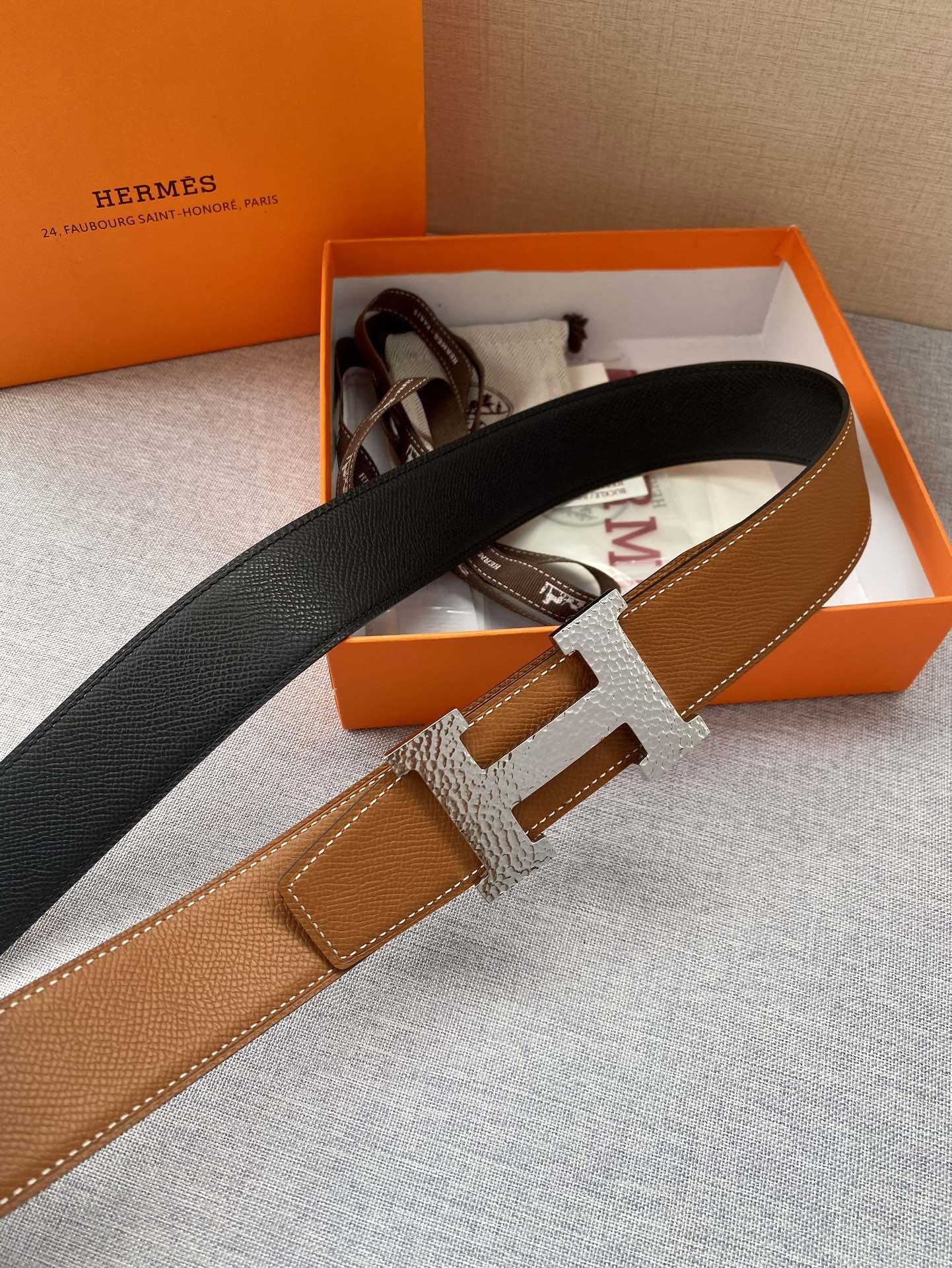 Tan leather belt with black strap back and gold-tone H buckle; side angle view.