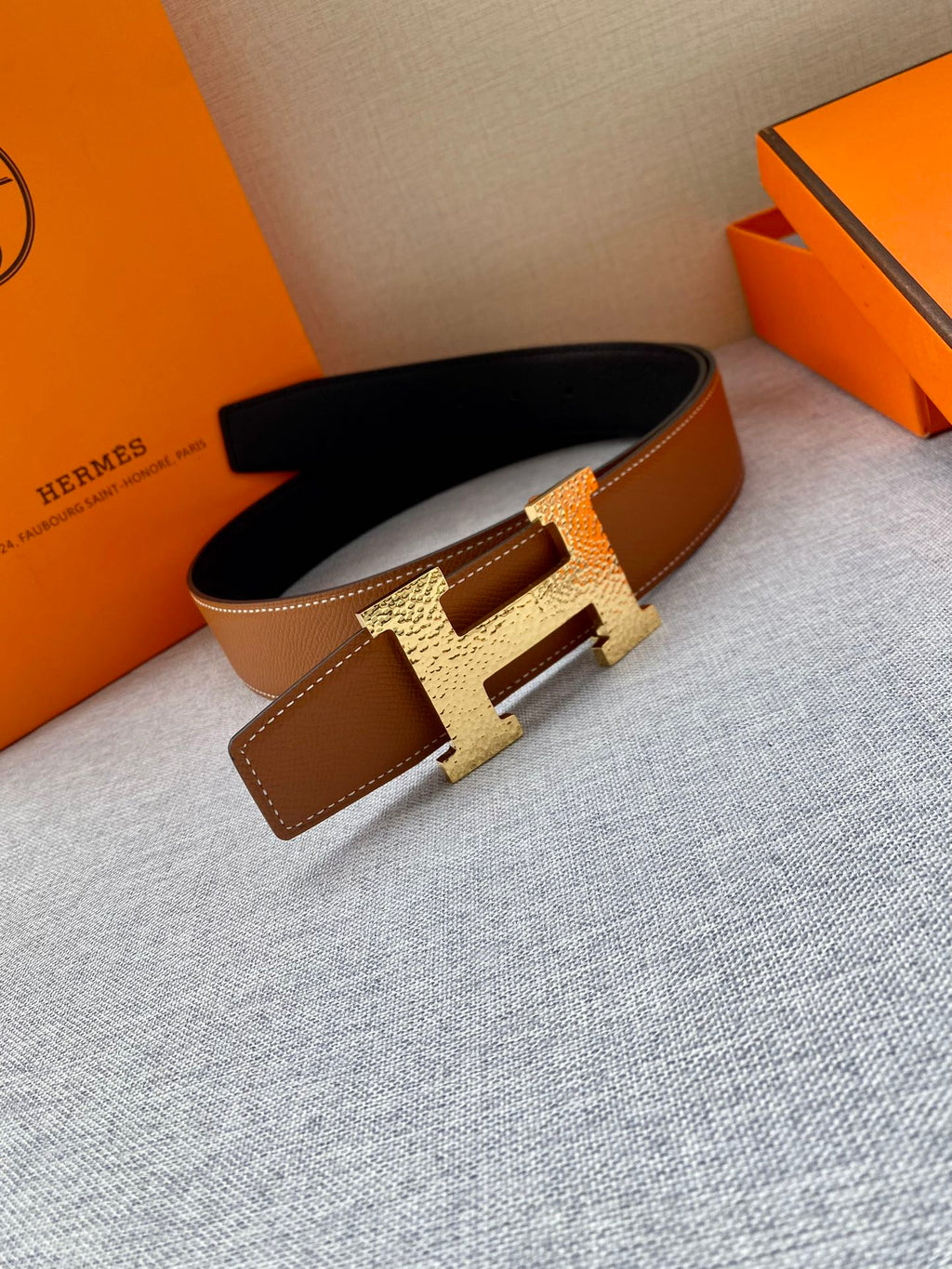 Tan leather belt with gold-tone H buckle; folded strap with keeper panel visible.