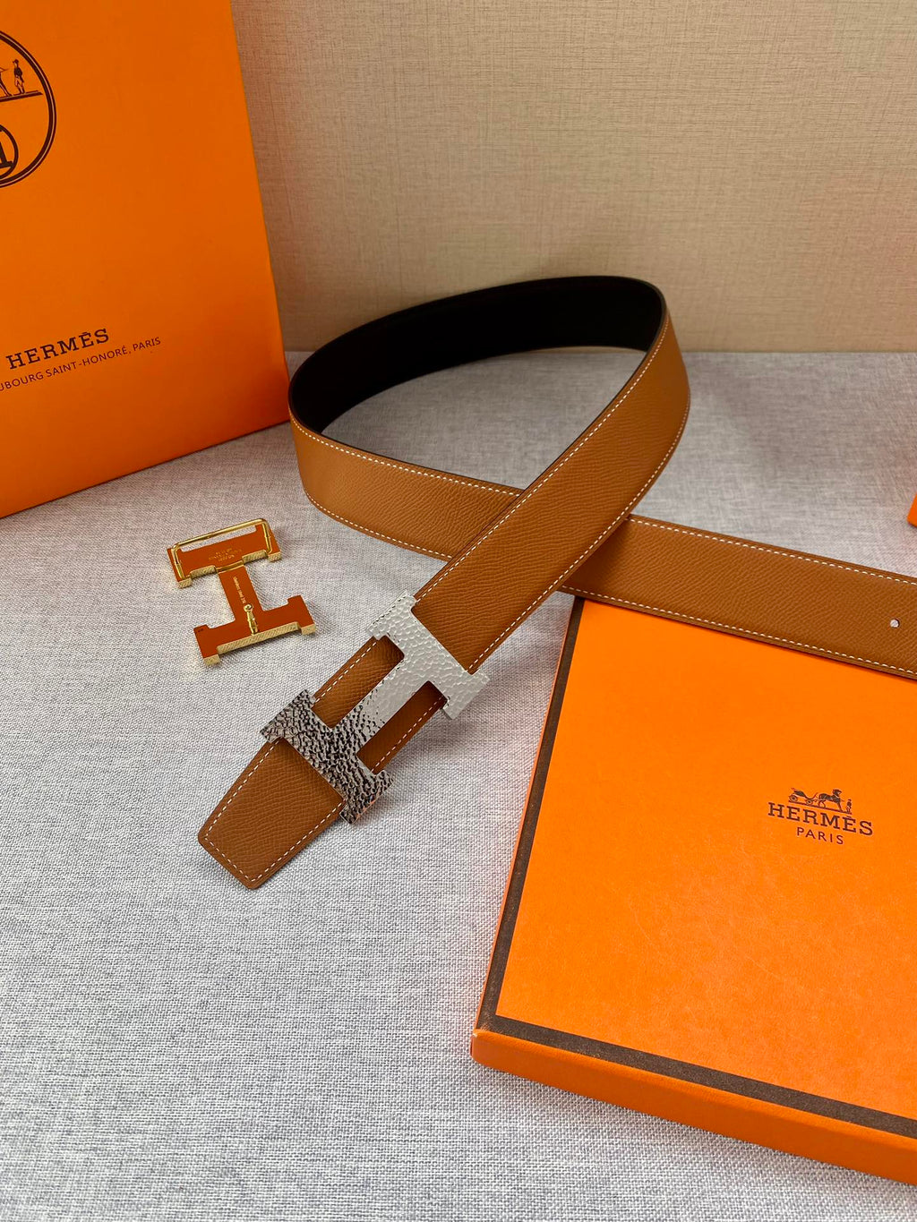 Tan and dark belt with gold-tone H buckle; belt laid beside the orange presentation box.