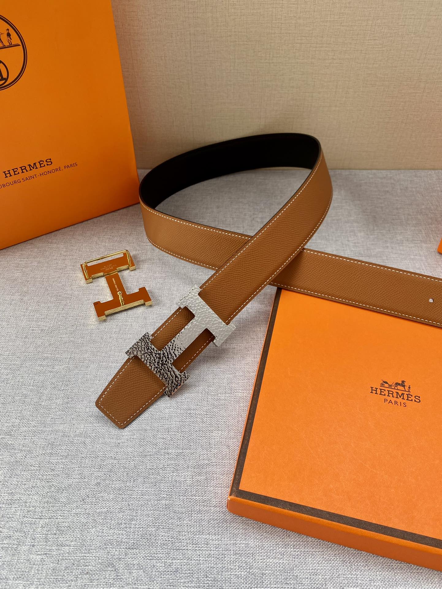 Tan and dark belt with gold-tone H buckle; belt laid beside the orange presentation box.