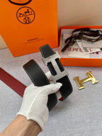 Hermes belt with black textured leather and gold-tone H buckle, shown at a diagonal angle.